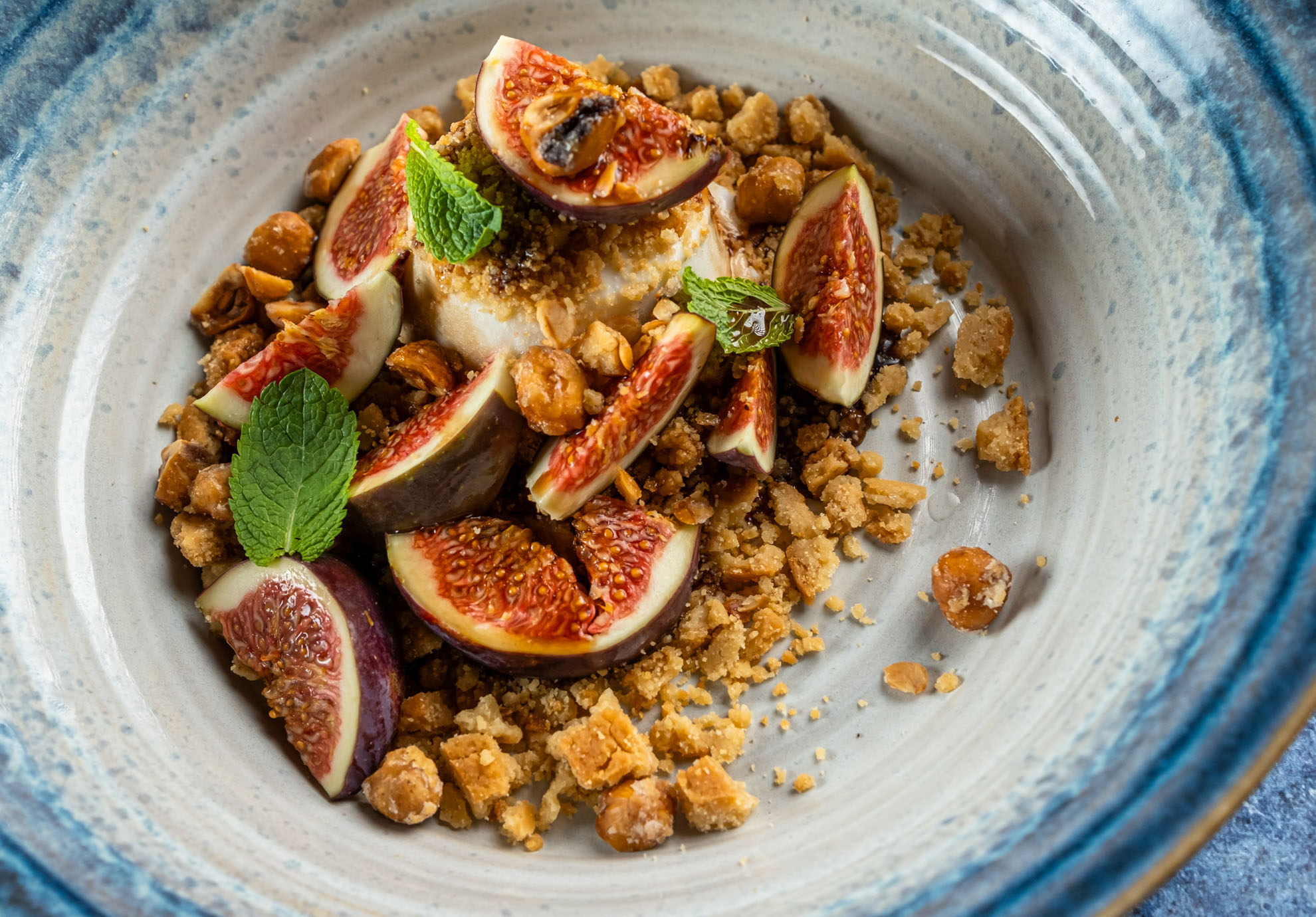 Ein Gourmetgericht mit cremiger Panna Cotta, garniert mit geschnittenen Feigen und Minzblättern auf einem beigen Teller, umgeben von zerbröselten Nüssen und Keksen.