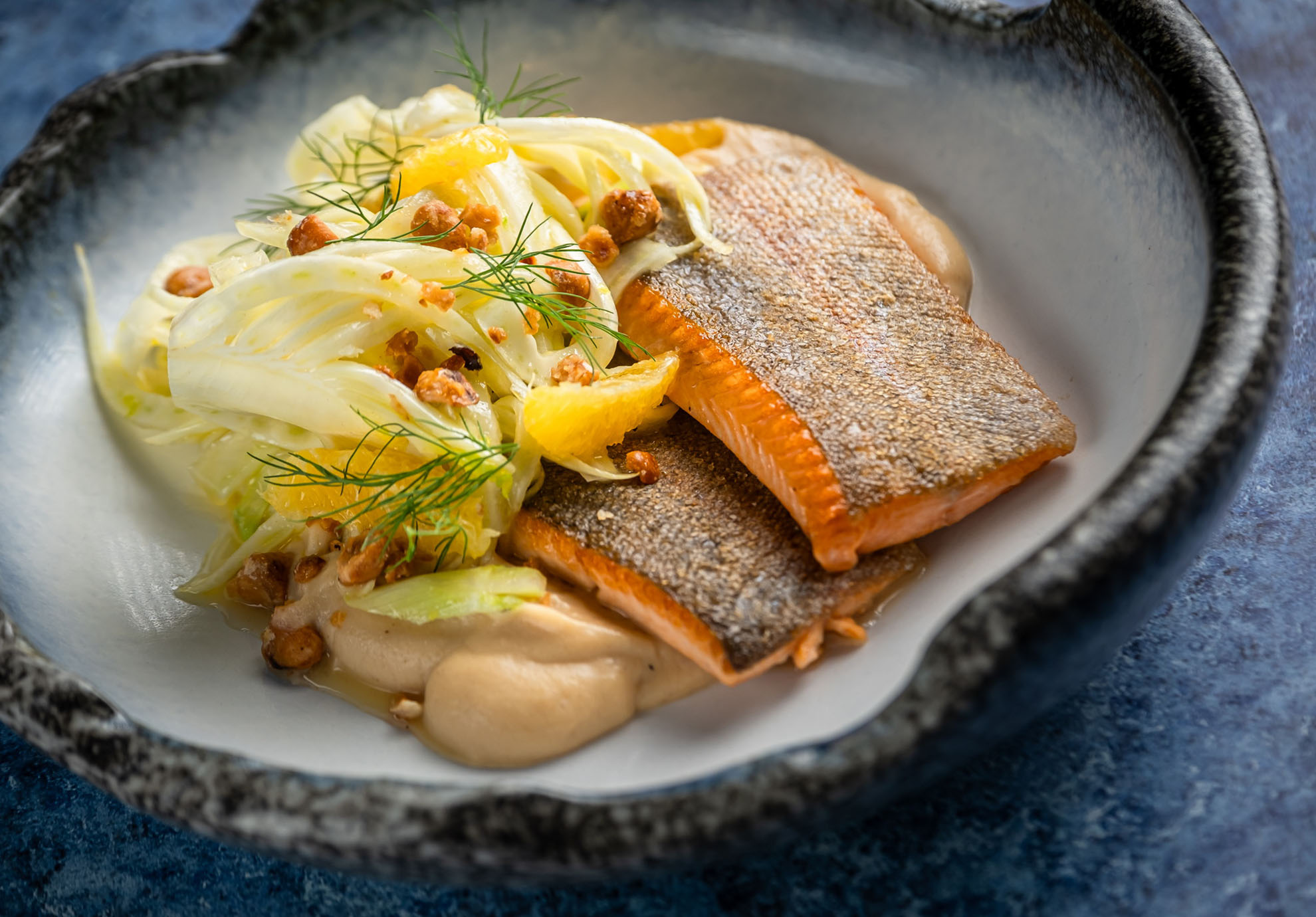 Ein Gericht mit gebratenem Lachs, knuspriger Haut nach oben, dazu gehobelter Fenchelsalat, Orangenschnitze, Nüsse und ein Klecks beiger Sauce in einer rustikalen Schüssel.