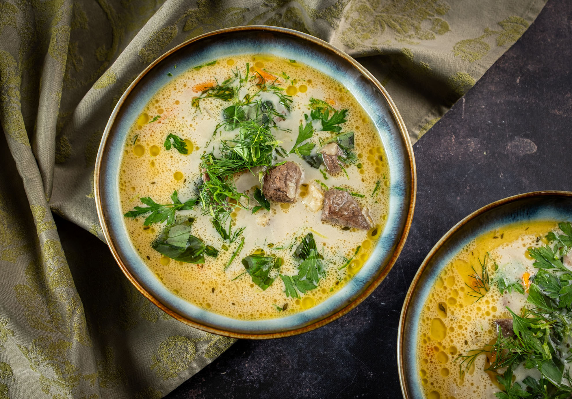 Eine Schüssel cremige Suppe mit Hirschfleischstücken, garniert mit frischen Kräutern wie Petersilie und Dill. Die Suppe steht auf einem strukturierten dunklen Untergrund neben einem gemusterten grünen Stoff.