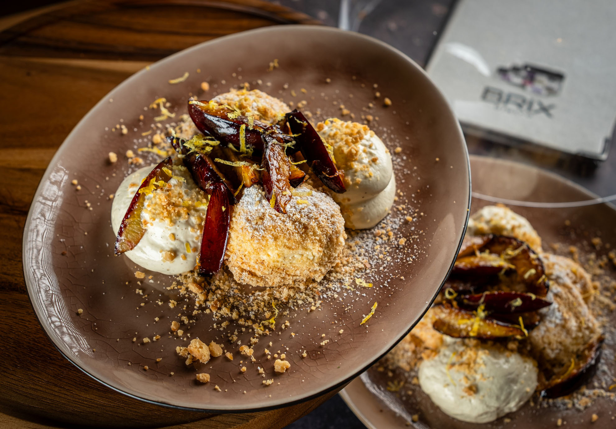 Ein rustikales Dessert auf einem braunen Teller, cremige Quarkknödel mit karamellisierten Feigen, Streuseln und Zitronenschale, serviert auf einem Holztisch.